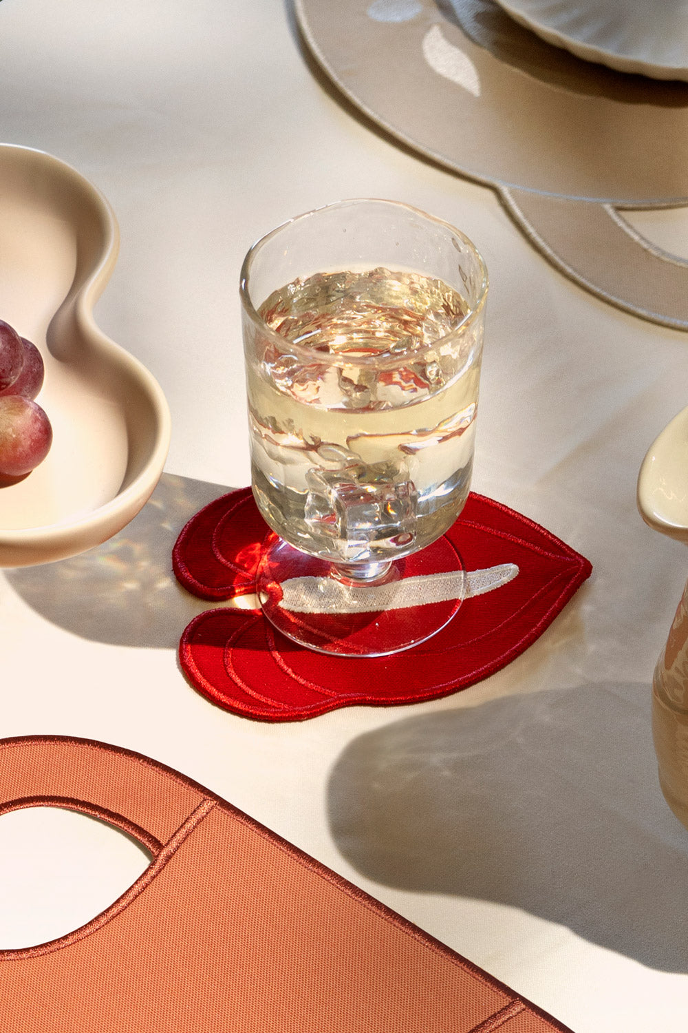 A glass of white wine rests on an Octaevo Red Anthurium Coaster beside plates and grapes on a cream tablecloth.