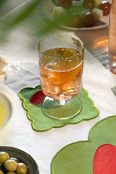 A glass filled with an amber drink sits on a green olive fabric coaster with a red accent, placed on a white tablecloth in a dining setting.