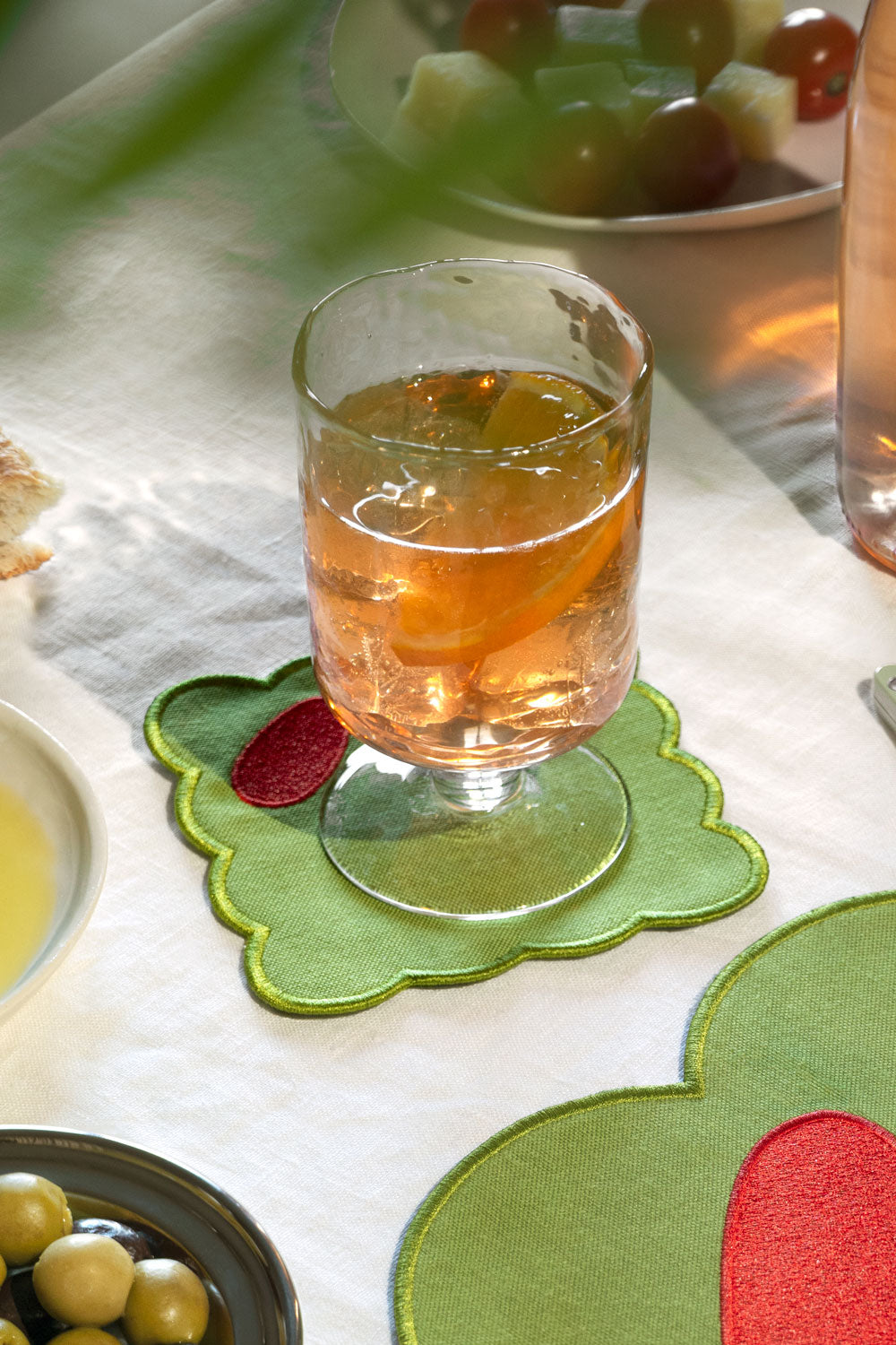 A glass filled with an amber drink sits on a green olive fabric coaster with a red accent, placed on a white tablecloth in a dining setting.