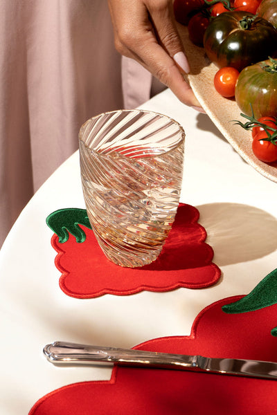 A hand sets a glass on an Octaevo Tomato Coaster (Set of 4) beside tomatoes and a knife on a white table.