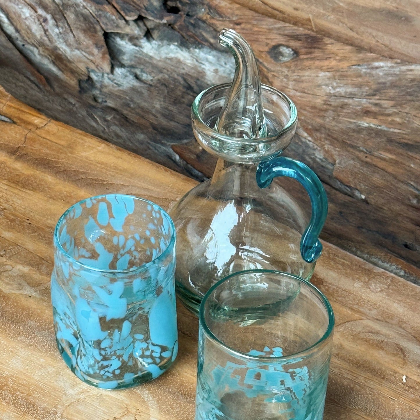 Two turquoise glass tumblers and a clear glass carafe with a blue handle on a wooden surface. - lifestyle