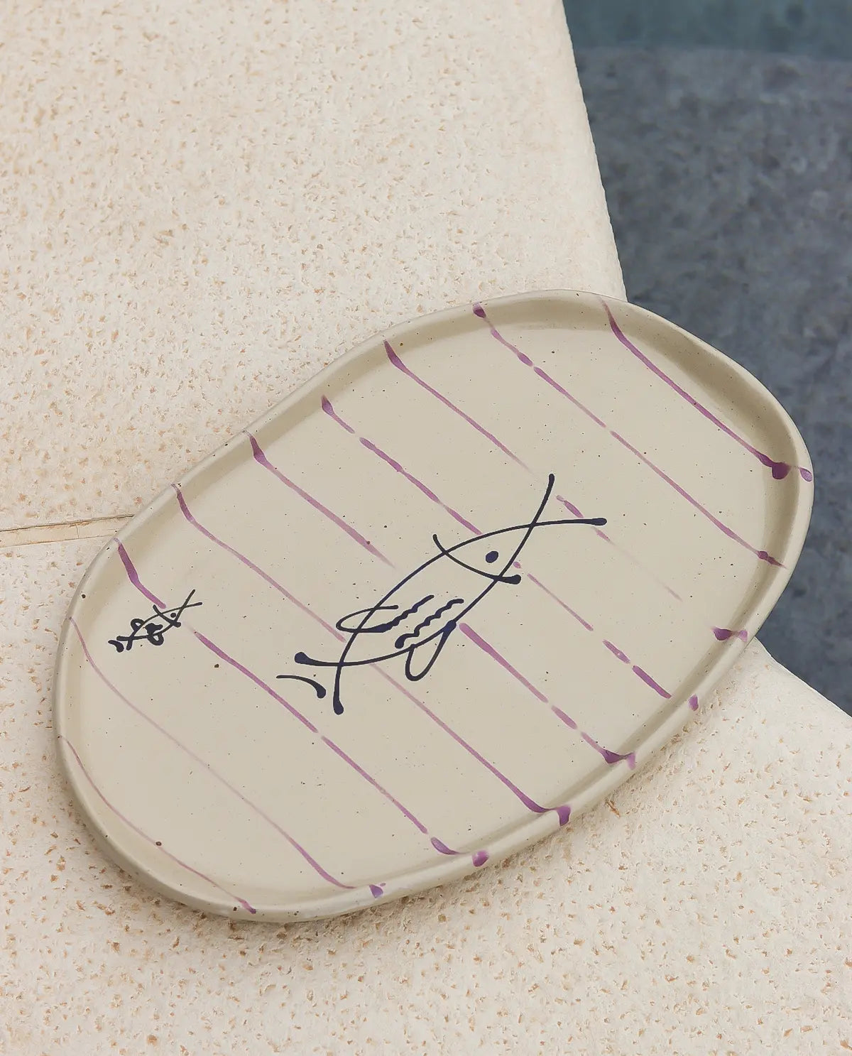 Oval ceramic plate with purple stripes and fish design on a textured surface