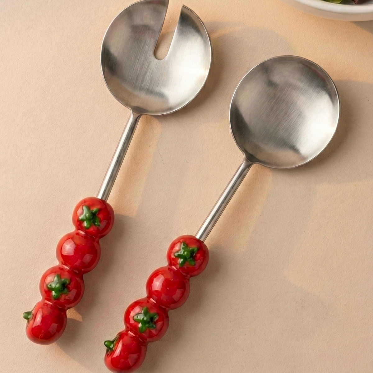 Two silver salad servers with tomato-themed handles on a beige surface, next to a bowl of salad.