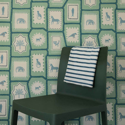 Folk Tales wallpaper in pistachio green with illustrated animal pattern shown in situ with chair for scale.