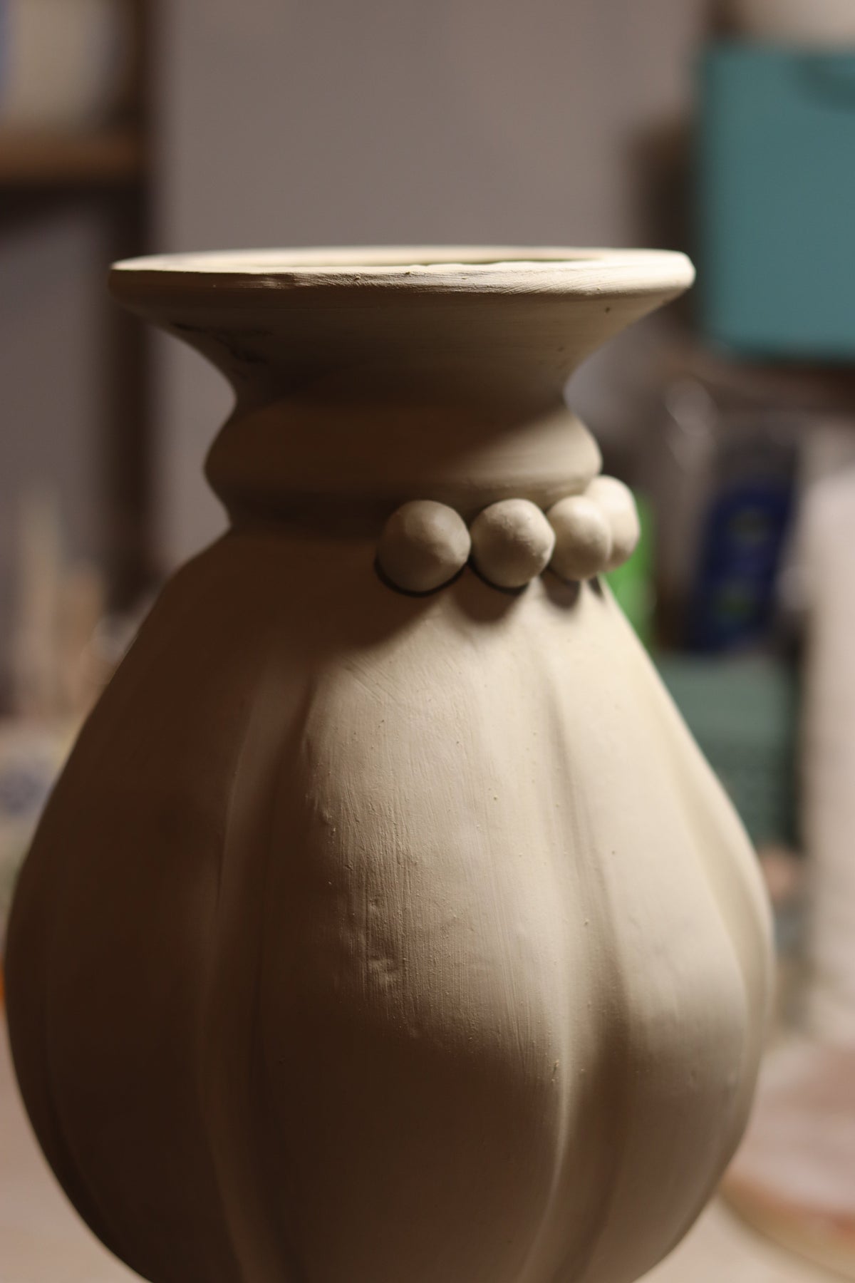 An unglazed ceramic vase in a light warm gray or beige clay color, shown in a close-up side profile against a blurred workshop background.