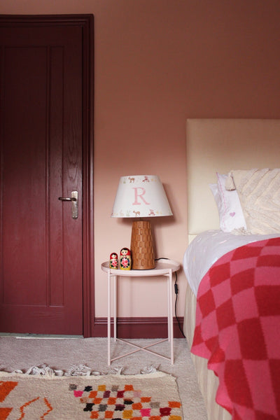 Personalised kids lampshade with woodland animal print and customised initial, styled on a ceramic lamp base in a cosy kids bedroom design with pink walls and playful décor.