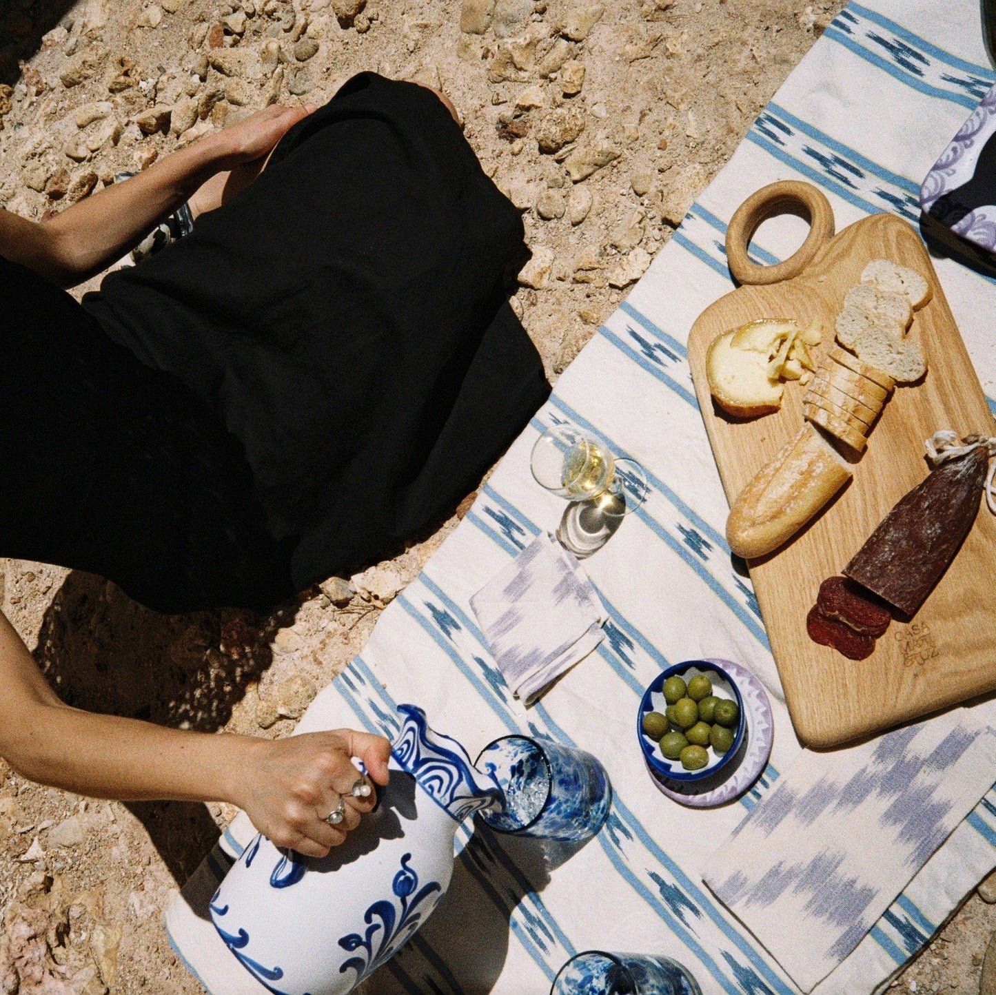 Person sitting on a rocky surface with a picnic setup including table runner, wood board and ceramic - lifestyle