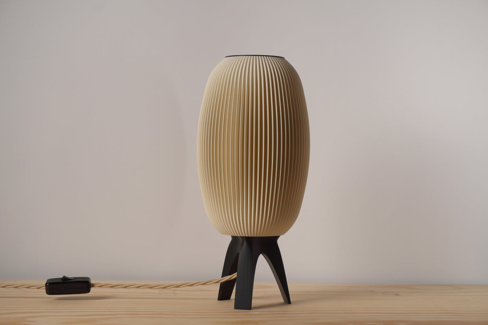 A beige, ribbed, mushroom-shaped table lamp with a tripod base, turned on, illuminating a warm light.