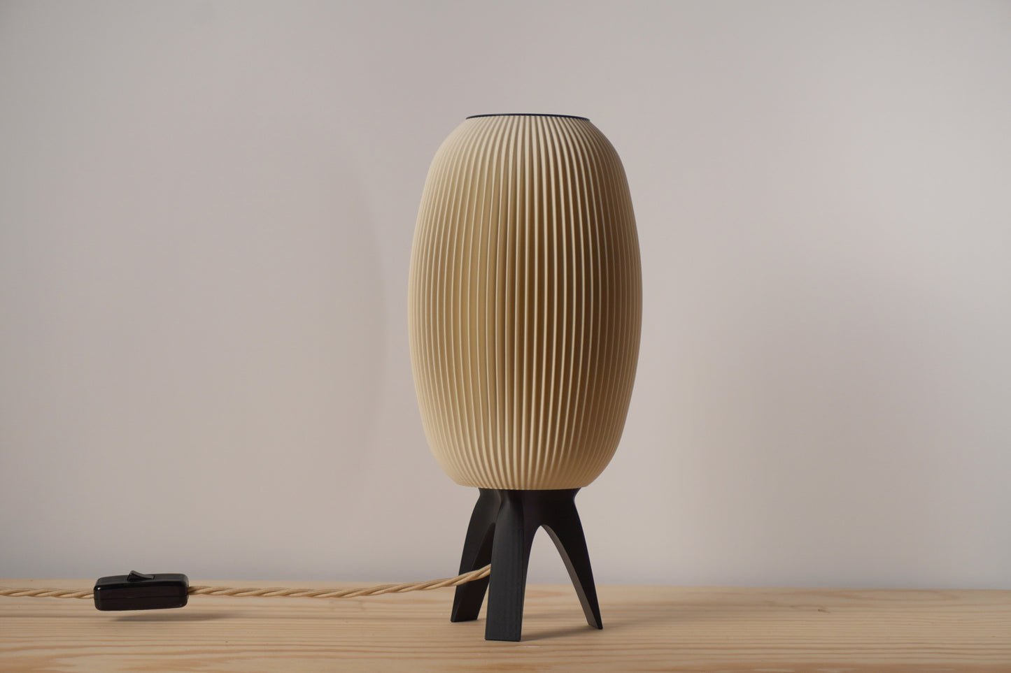 A beige, ribbed, mushroom-shaped table lamp with a tripod base, turned on, illuminating a warm light.