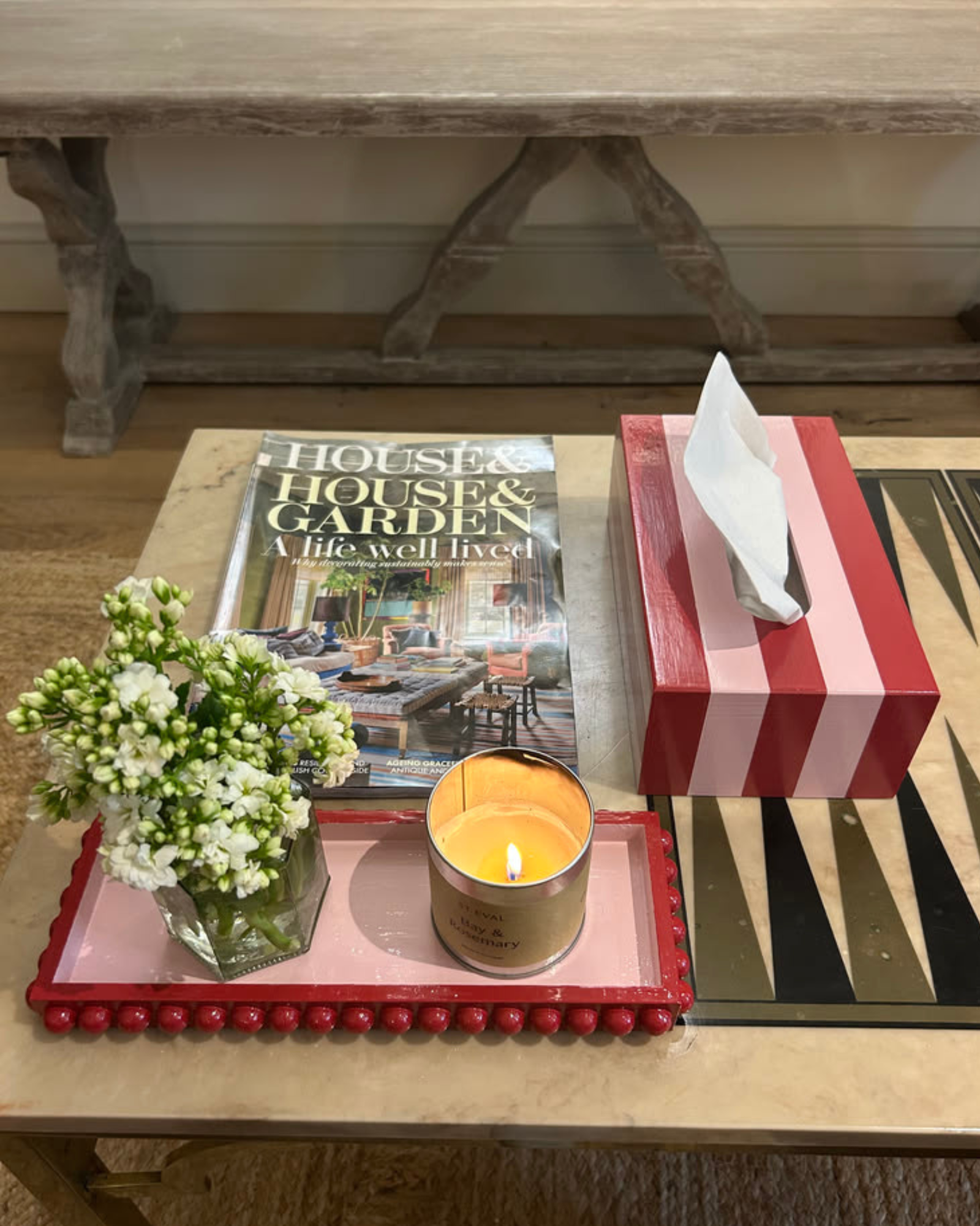 A styled tabletop scene with a red-and-white striped tissue box, a pink tray with a small flower arrangement and a lit candle, a magazine, and a black-and-gold geometric mat, set in a home interior.