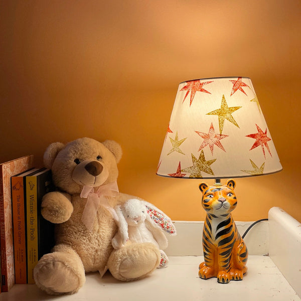 A dimly lit kids bedroom corner with a cream lampshade decorated with bright orange and yellow star patterns, styled in a kids bedroom on a tiger lamp base. A fun empire lampshade and kids bedroom decor piece that adds warmth and character.