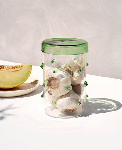 The OCTAEVO Agave Glass Container Large with a green lid holds garlic; a melon slice sits on a nearby plate.