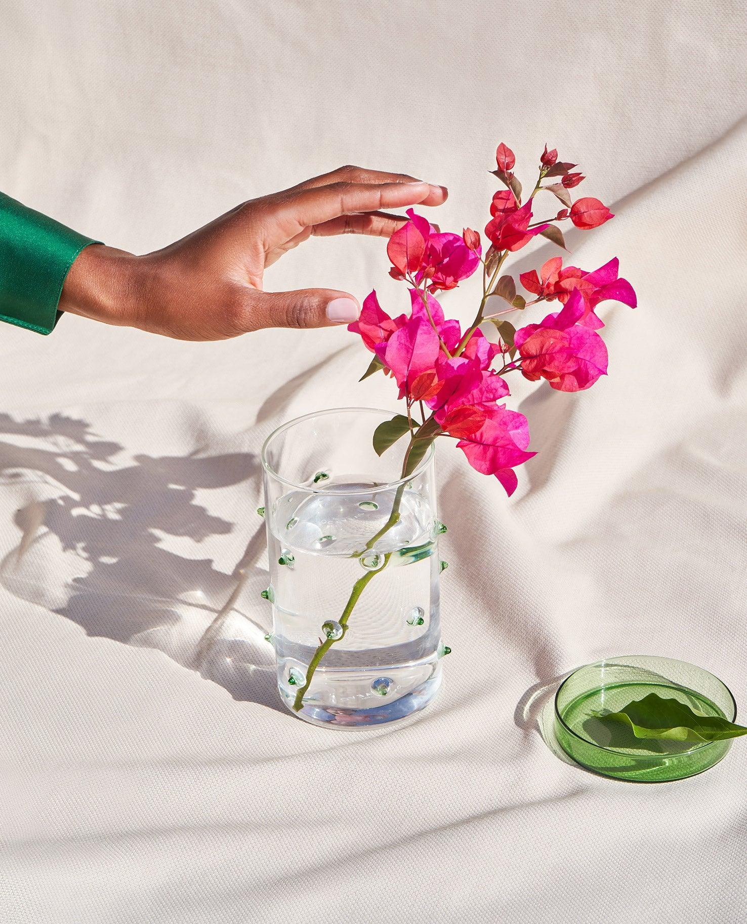 A hand reaches for the OCTAEVO Agave Glass Container Large beside pink flowers on a light fabric background.