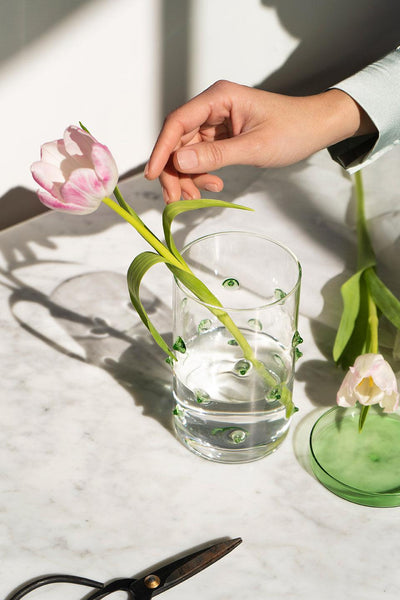 A hand places a pink tulip in the Agave Glass Container Large by OCTAEVO on a marble table, scissors nearby.