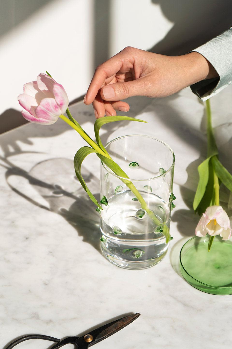 A hand places a pink tulip in the Agave Glass Container Large by OCTAEVO on a marble table, scissors nearby.