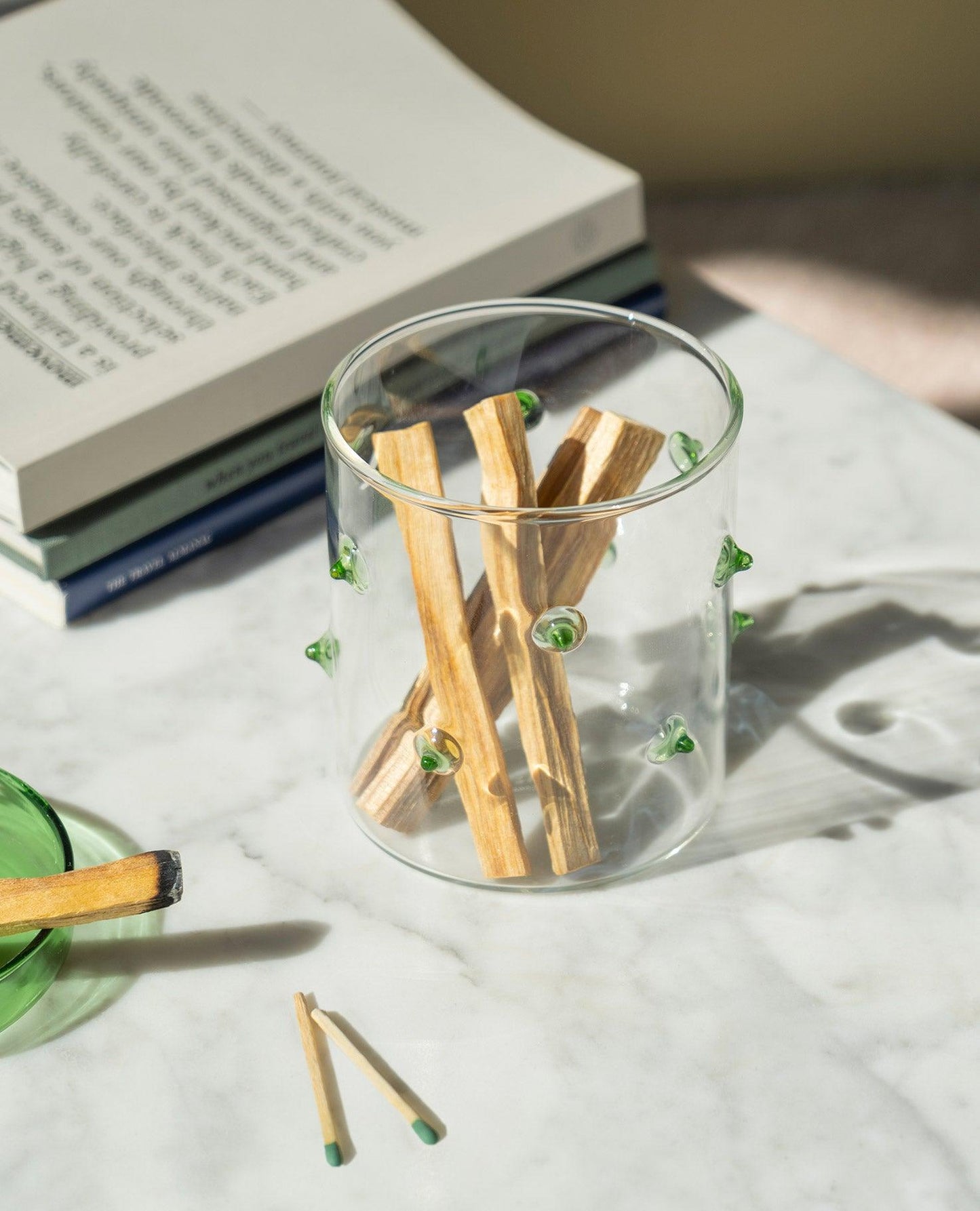 The Agave Glass Container Small by OCTAEVO with green dots holds wooden sticks on a sunlit marble surface.