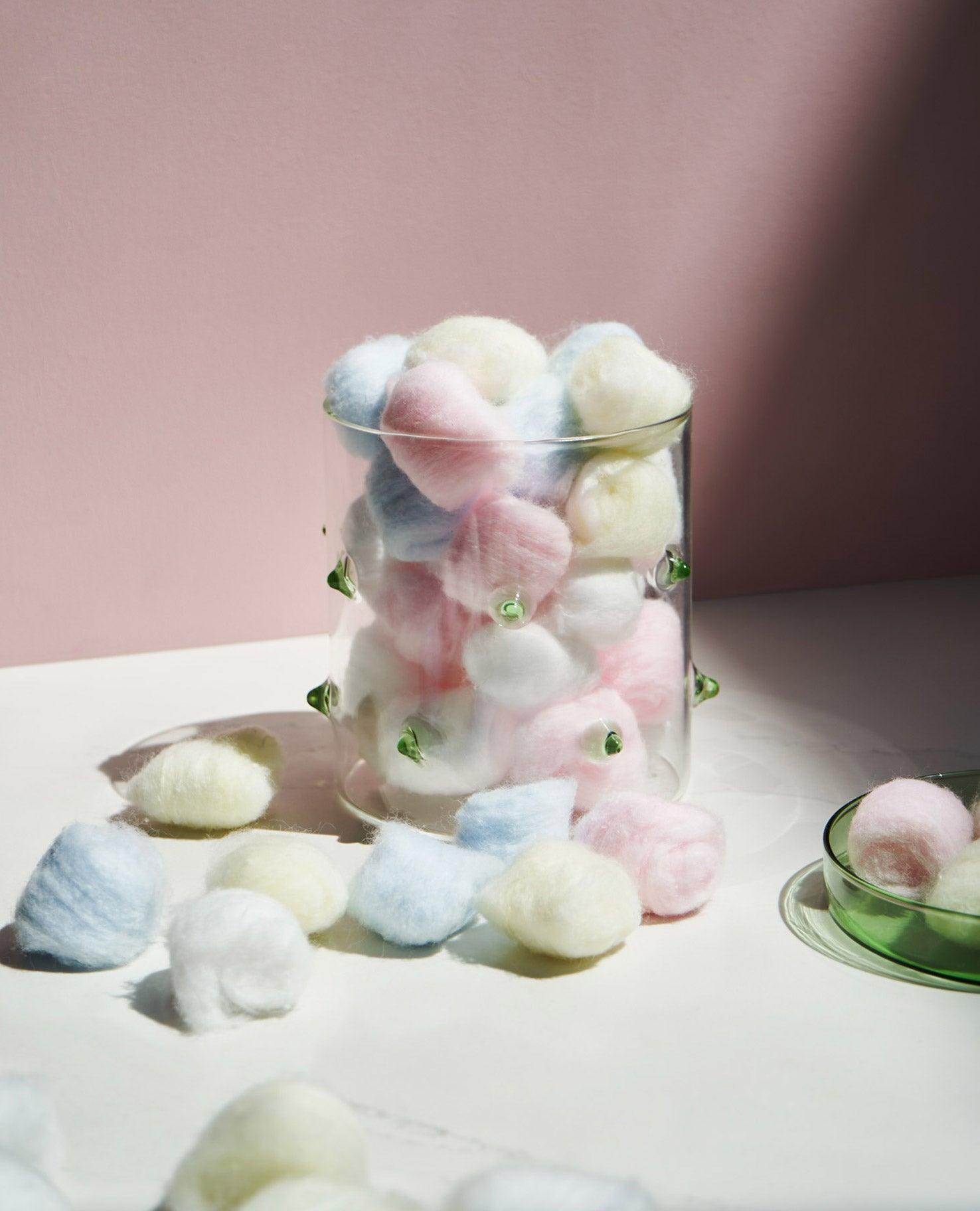 The OCTAEVO Agave Glass Container Small, filled with pastel cotton balls, sits beside a green dish and scattered balls.