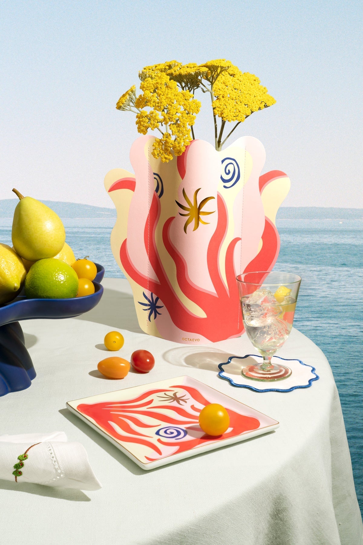 A colorful table setting with fruit, a floral vase, a glass of water, and a plate, set against a seaside background.