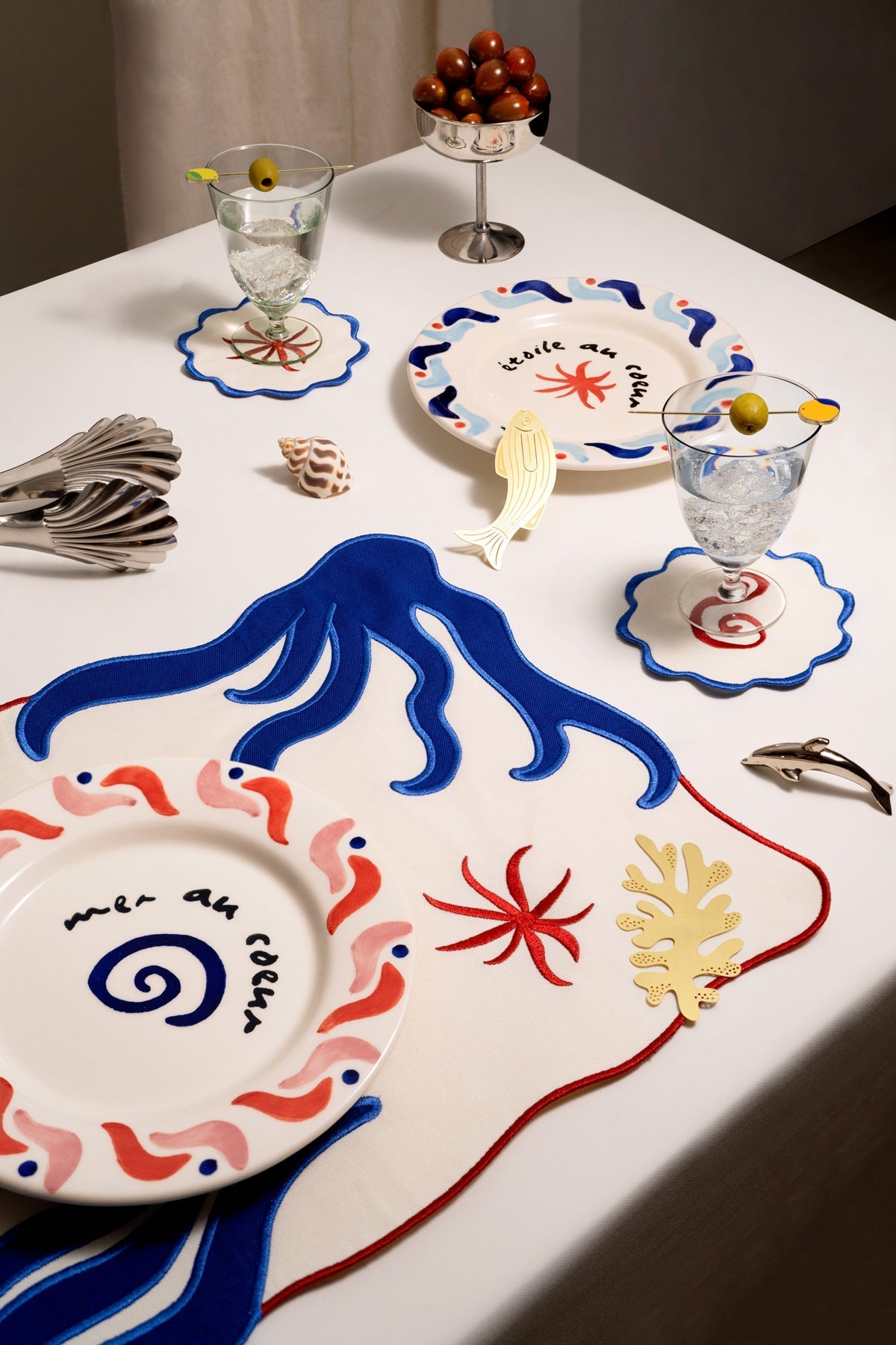 A table set with ocean plates, OCTAEVO Amalfi Placemats (Set of 2), olive glasses, and seashell decor.