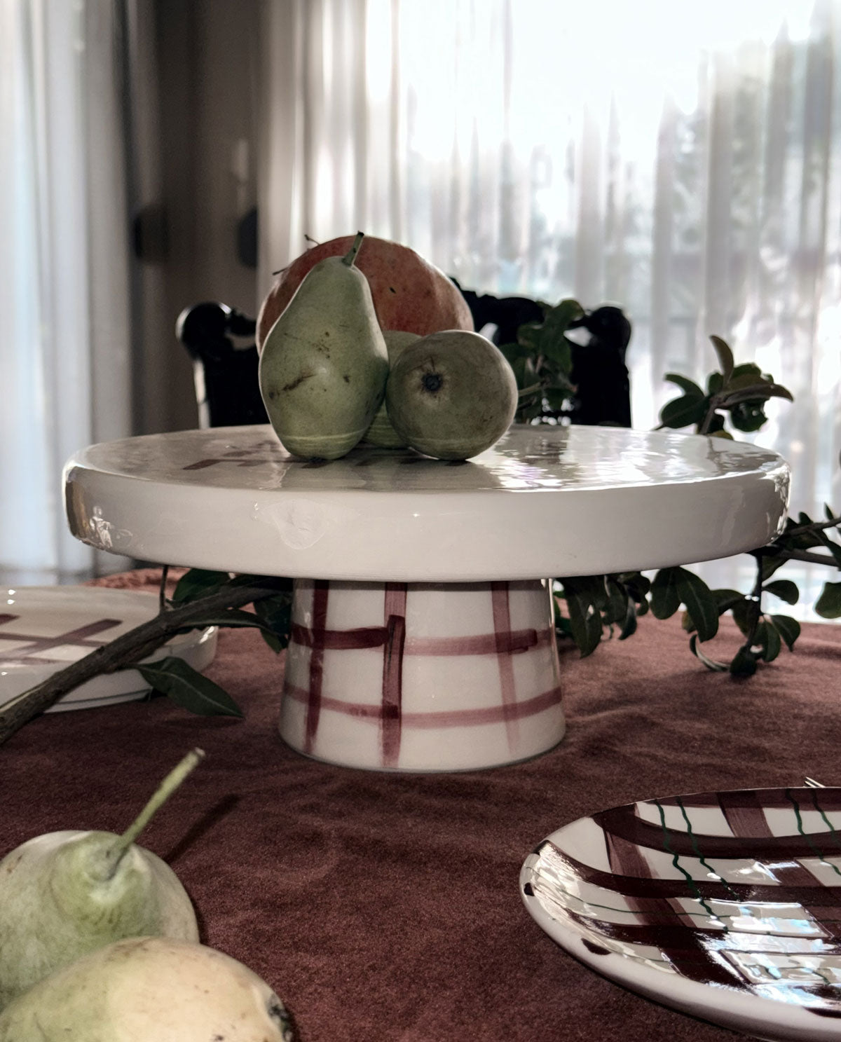 A round white ceramic cake stand with a red plaid pattern, styled on a dark tablecloth with pears and a peach on top, in a softly lit indoor setting.