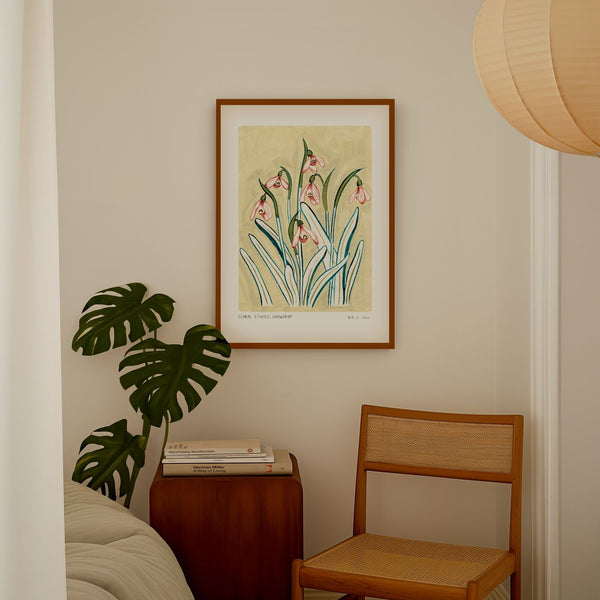 Snowdrop art print, a light muted yellow painted background with pinky snowdrop flowers and olive green/ turquoise leaves. With small text reading floral studies: snowdrop no.2 styled in a dark wood frame in a bedroom