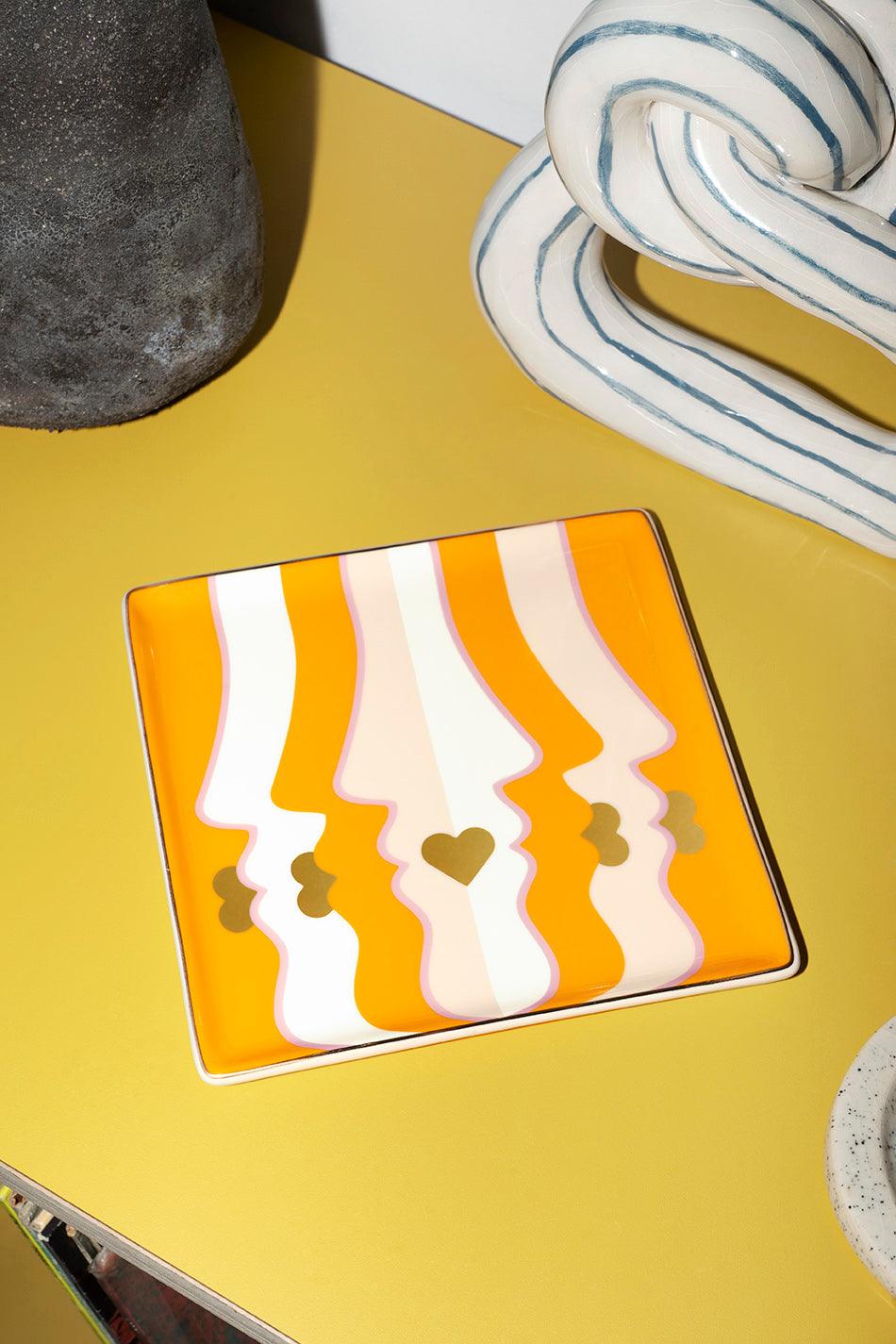 The orange Beso Ceramic Tray by Octaevo features abstract faces and a heart, adding vibrant style to any table.