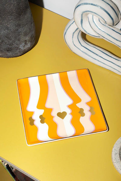 The orange Beso Ceramic Tray by Octaevo features abstract faces and a heart, adding vibrant style to any table.