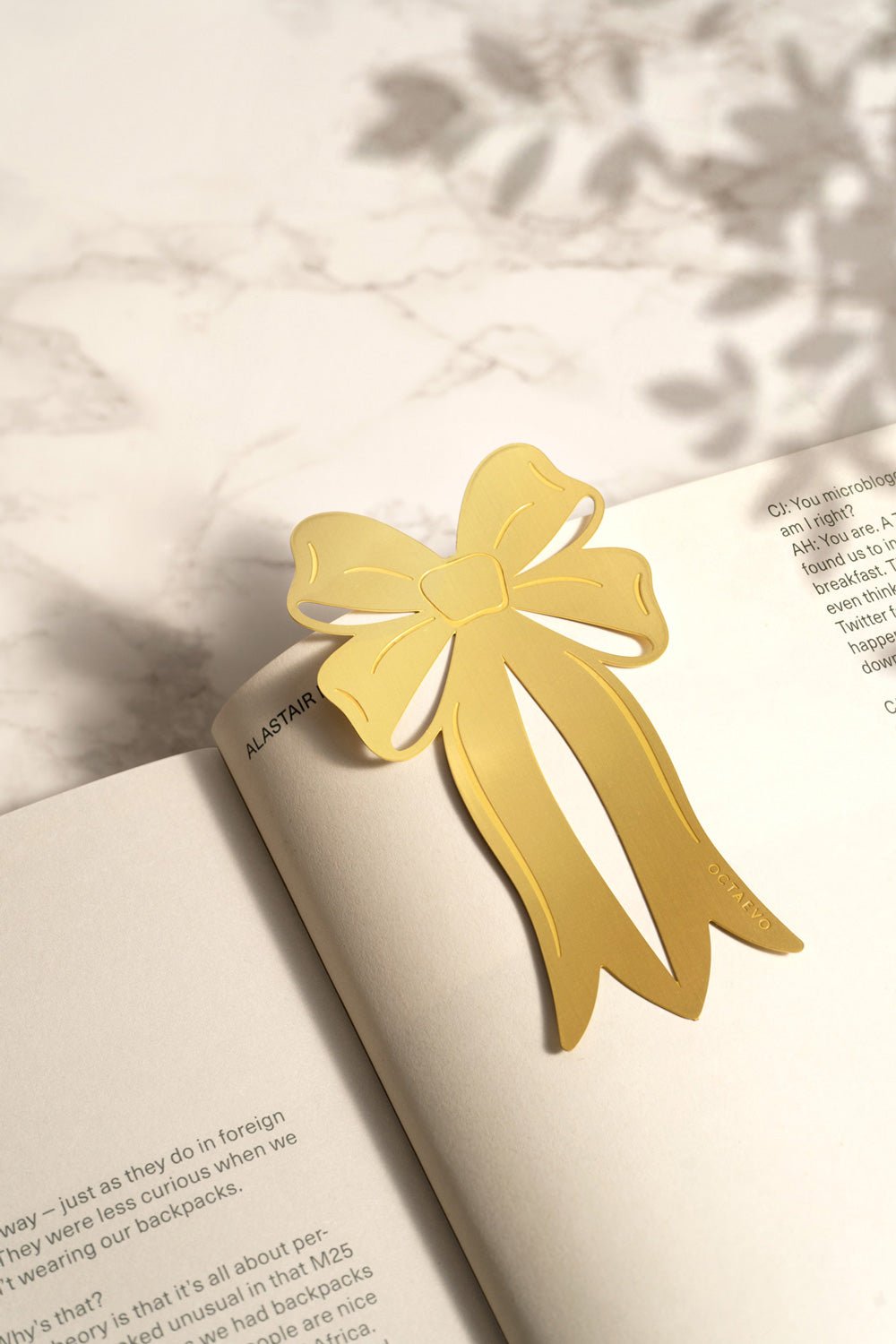 The OCTAEVO Bookmark Coquette, ribbon-shaped, rests on an open book, casting a soft shadow on marble.