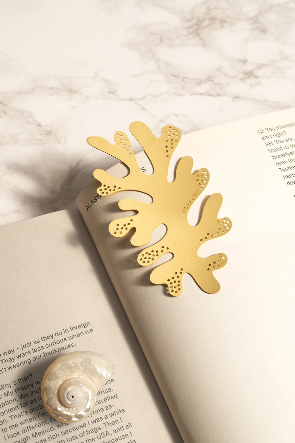 The OCTAEVO Bookmark Coral, a luxurious brass accessory, rests on an open book beside a seashell on marble.