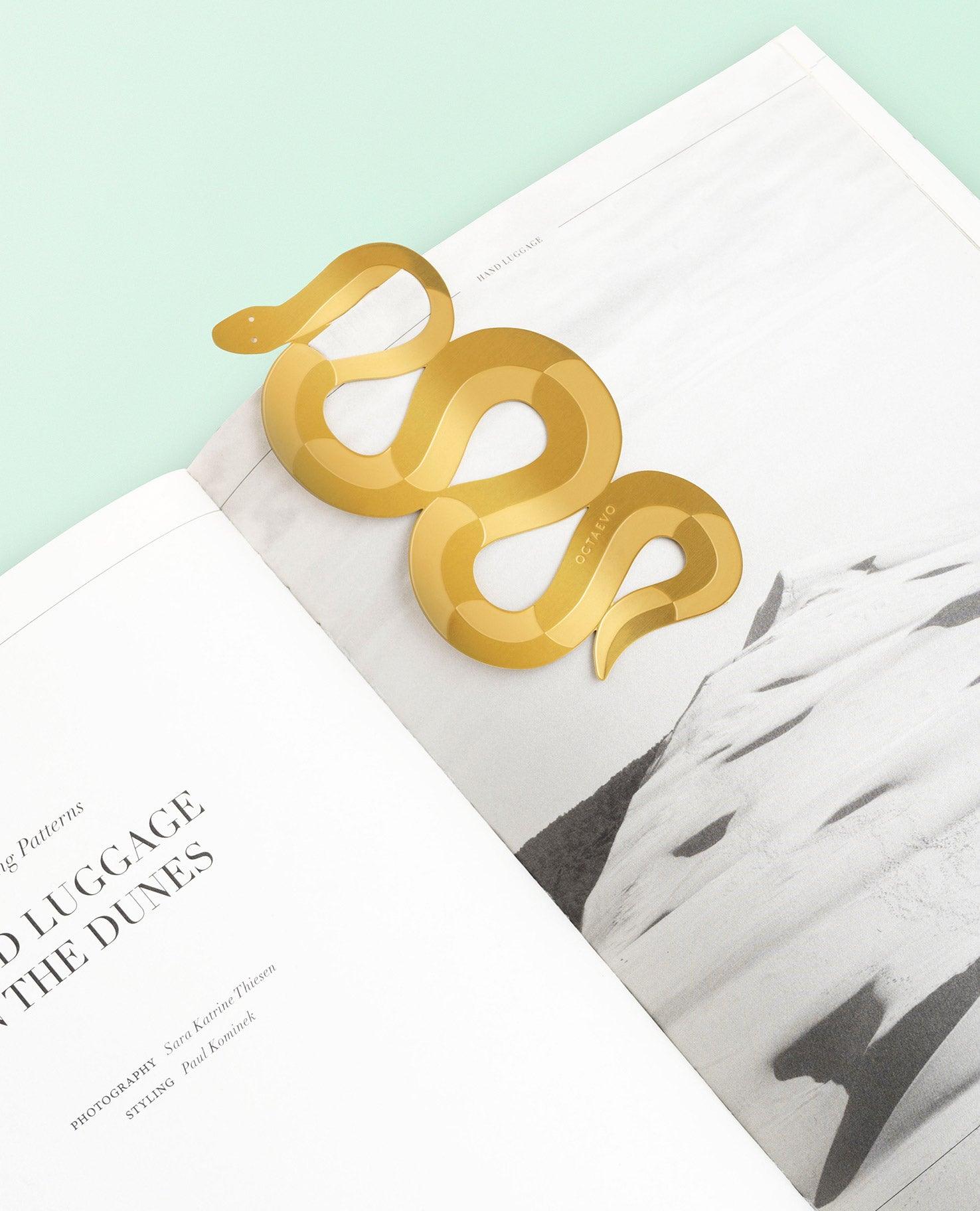 The OCTAEVO Bookmark Serpenti, shaped like a brass serpent, rests on an open book with a landscape photo.