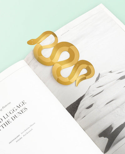 The OCTAEVO Bookmark Serpenti, shaped like a brass serpent, rests on an open book with a landscape photo.