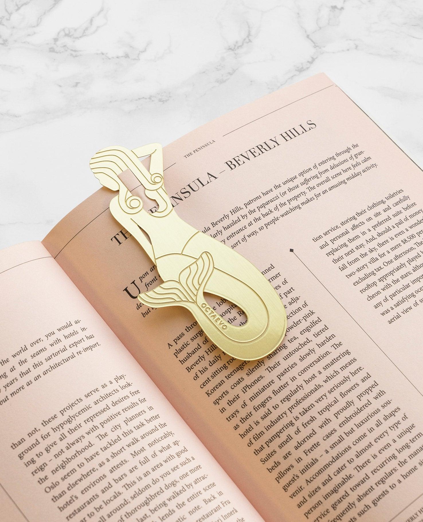 The OCTAEVO Bookmark Sirena, a gold brass abstract mermaid shape, rests on an open book with beige pages. 