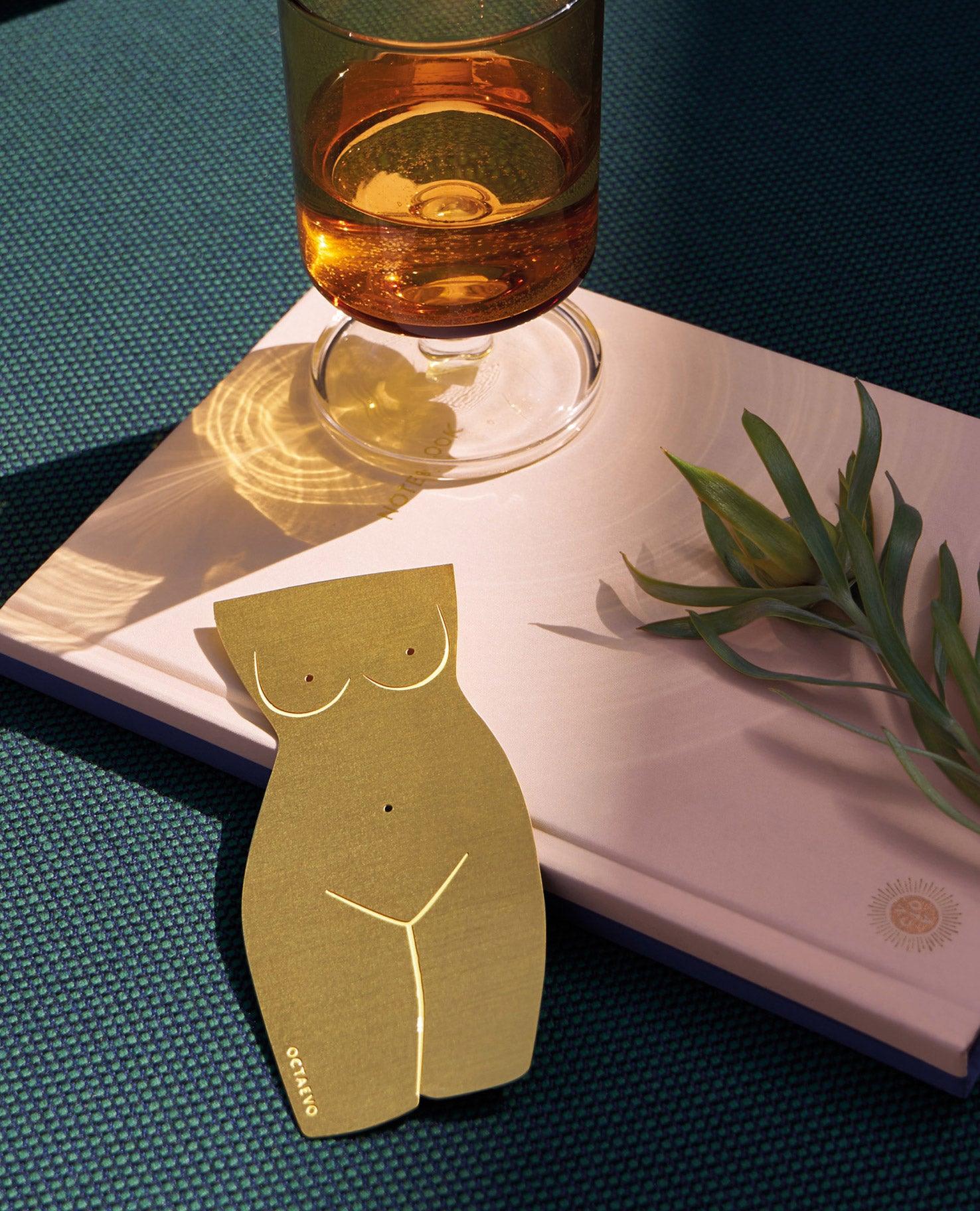The brass OCTAEVO Bookmark Venus rests atop a pink book with amber glass and greenery on a green surface.