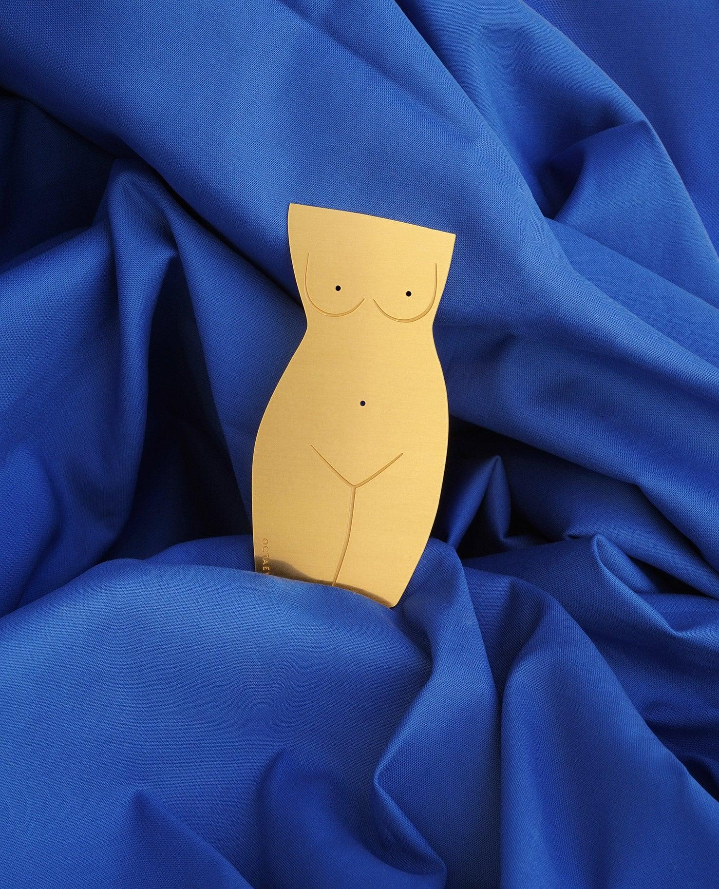 The OCTAEVO brass Bookmark Venus, a finely-cut gold female body sculpture, rests on royal blue fabric.