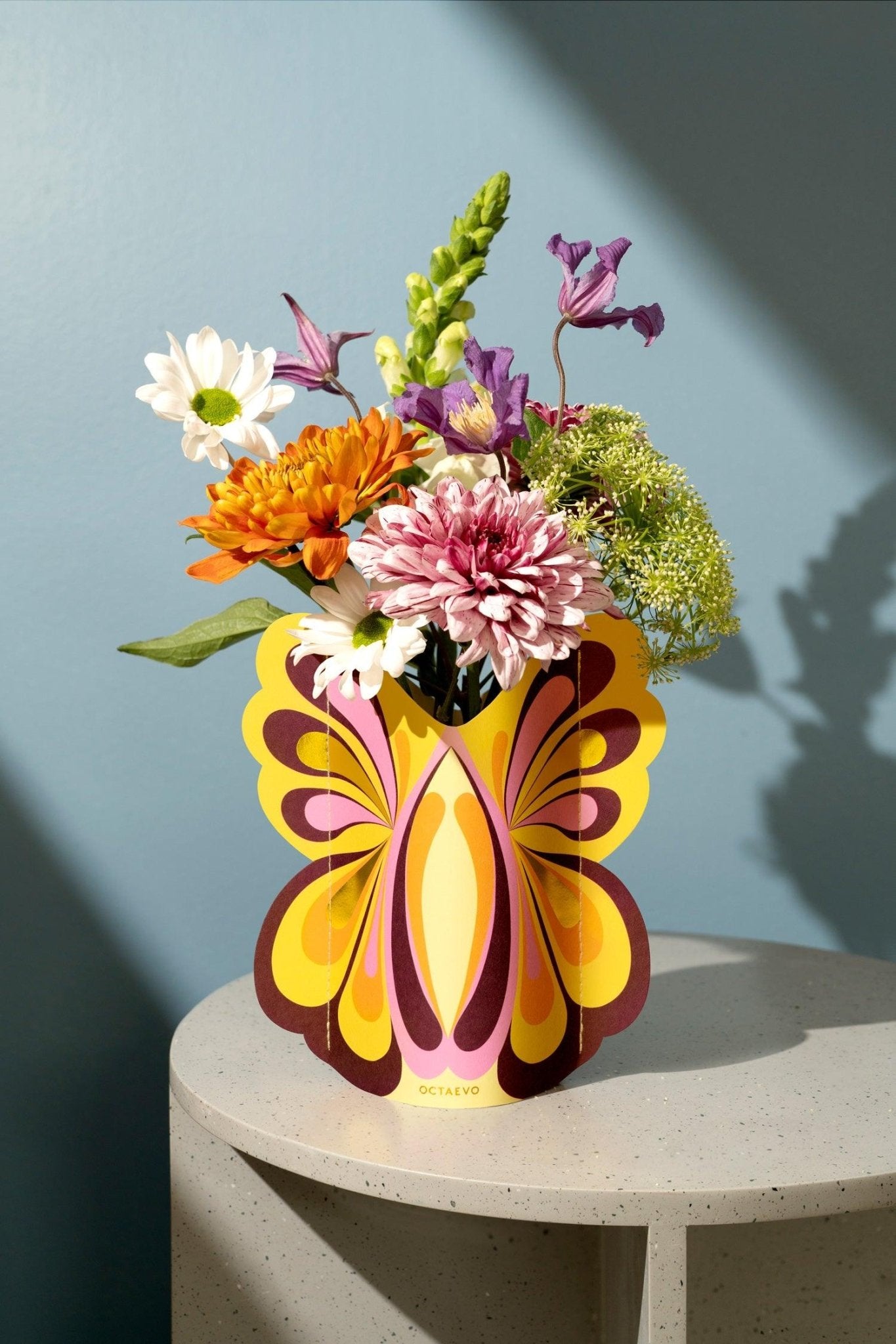 A vibrant bouquet in Octaevos Butterfly Mini Paper Vase rests on a speckled table against a blue wall.