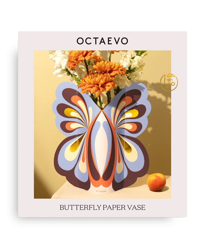 The Octaevo Butterfly Paper Vase, colorful and handcrafted, holds orange flowers with an orange fruit beside it.
