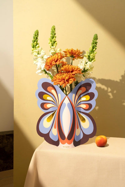 The Octaevo Butterfly Paper Vase holds flowers on a beige table, with a peach beside it.