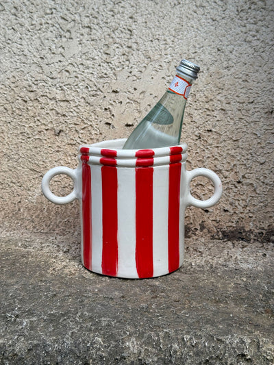 A ceramic wine cooler with red and white stripes, featuring two handles and a bottle inside, placed on a textured surface.