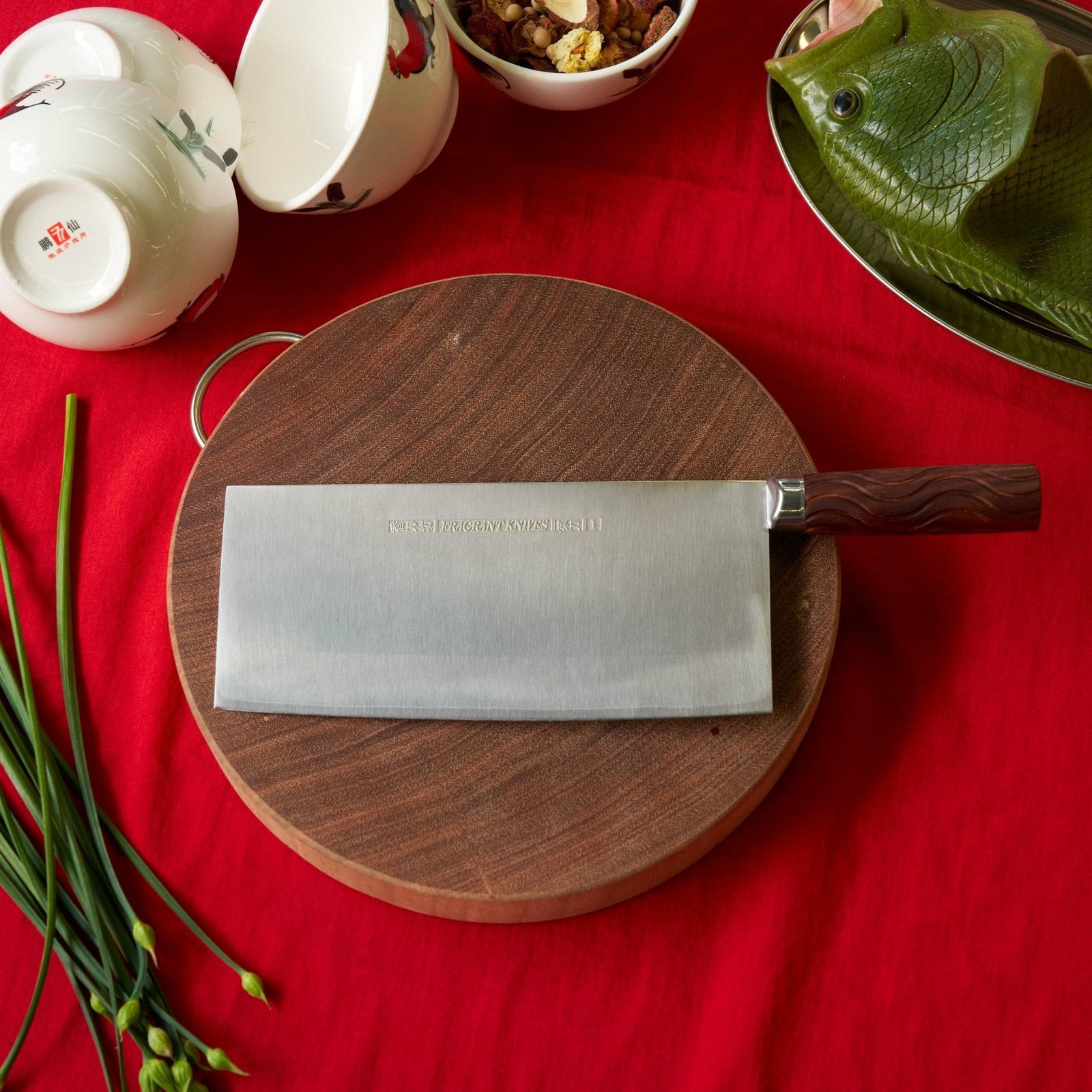 Wooden cutting board with a cleaver on a red tablecloth, surrounded by kitchen items.