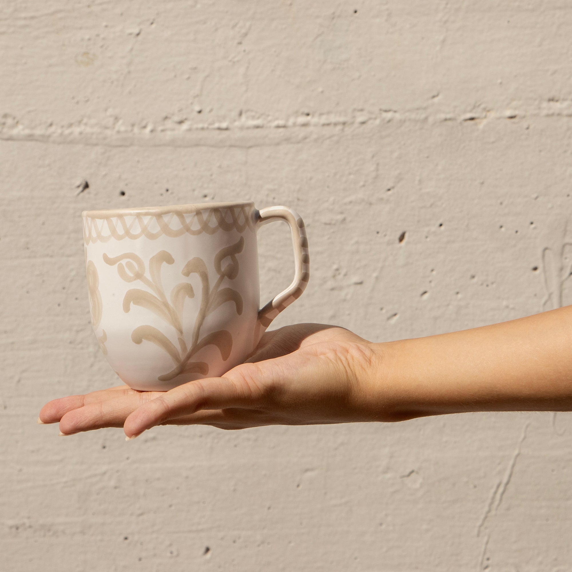 Hand holding a ceramic mug with decorative patterns against a beige wall. - lifestyle