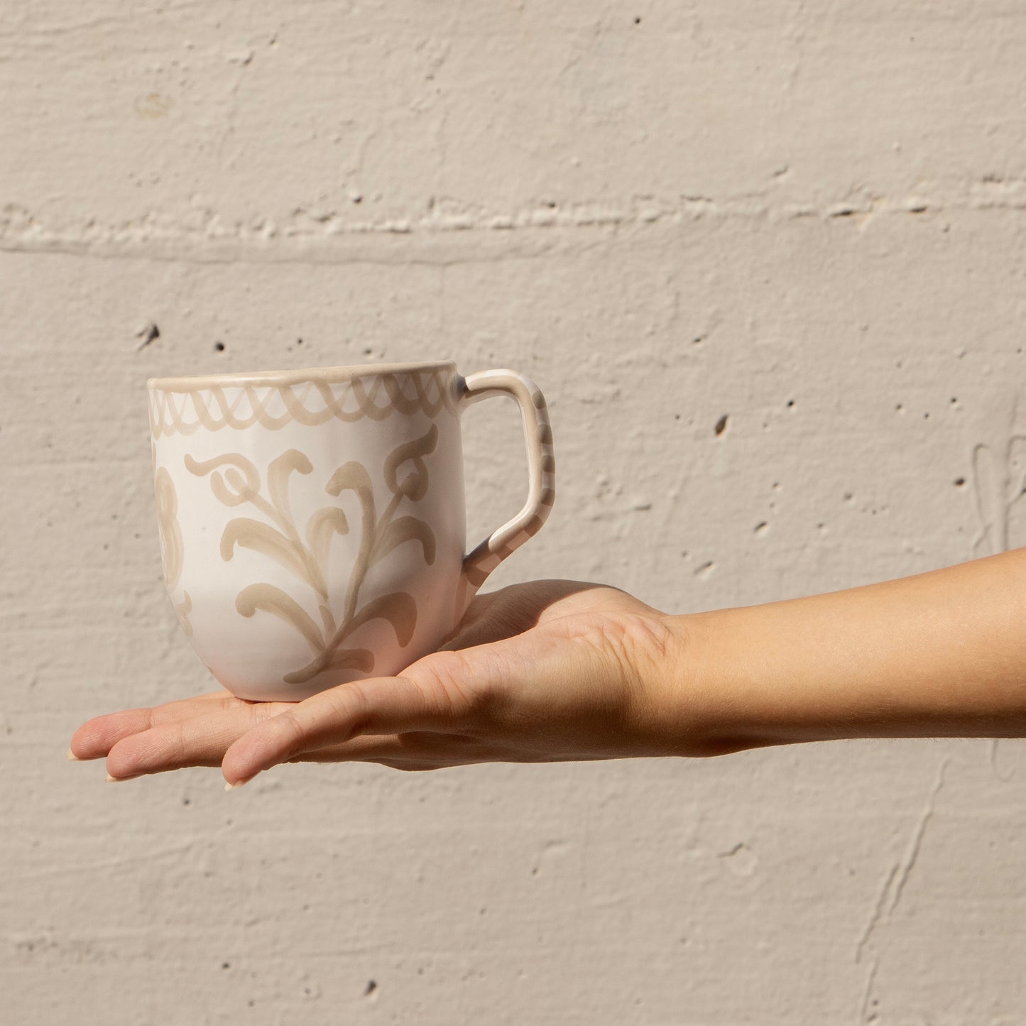 Hand holding a ceramic mug with decorative patterns against a beige wall. - lifestyle