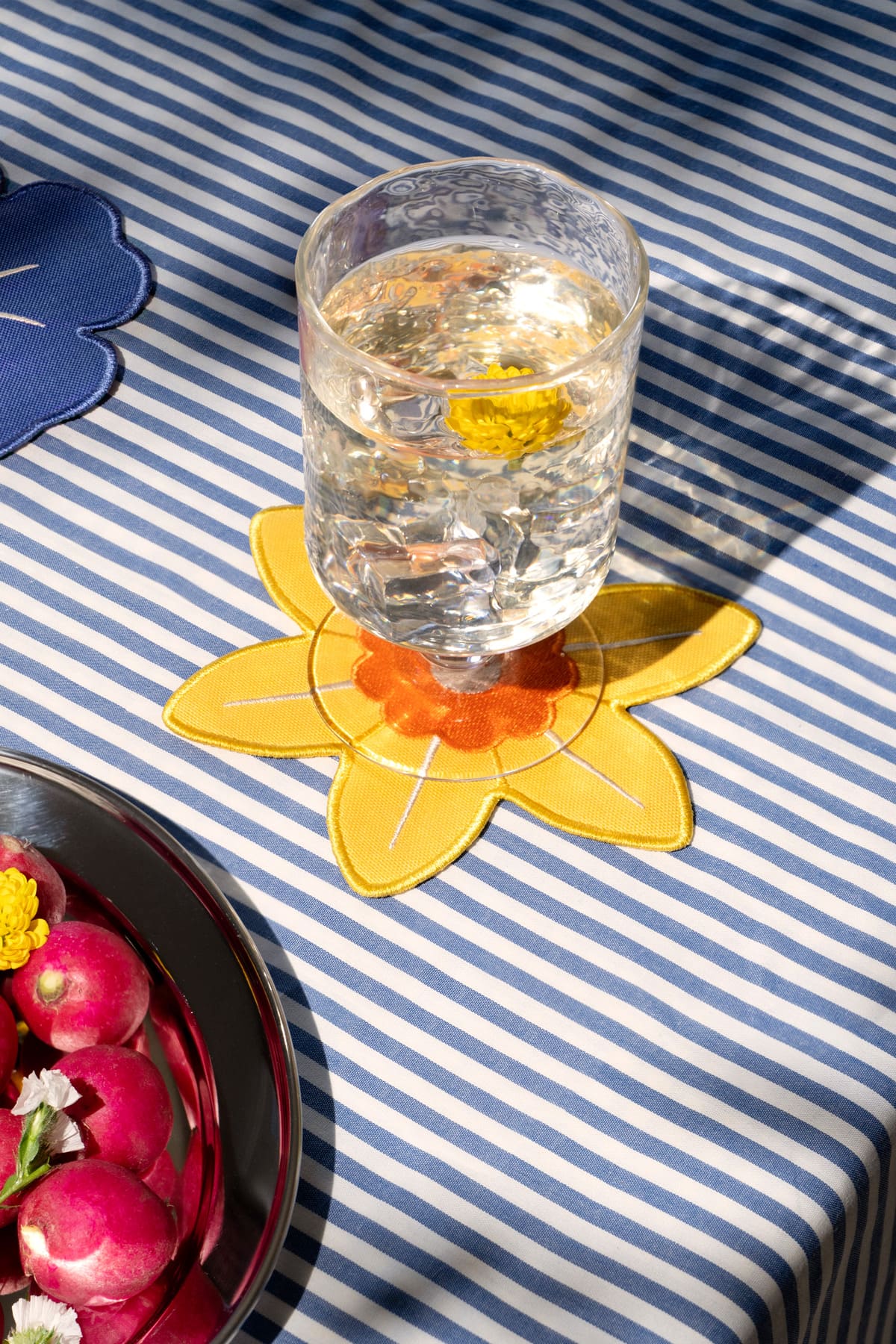 A glass of water with a yellow flower rests on Octaevo Daffodil Coasters (Set of 4) atop a blue striped tablecloth.