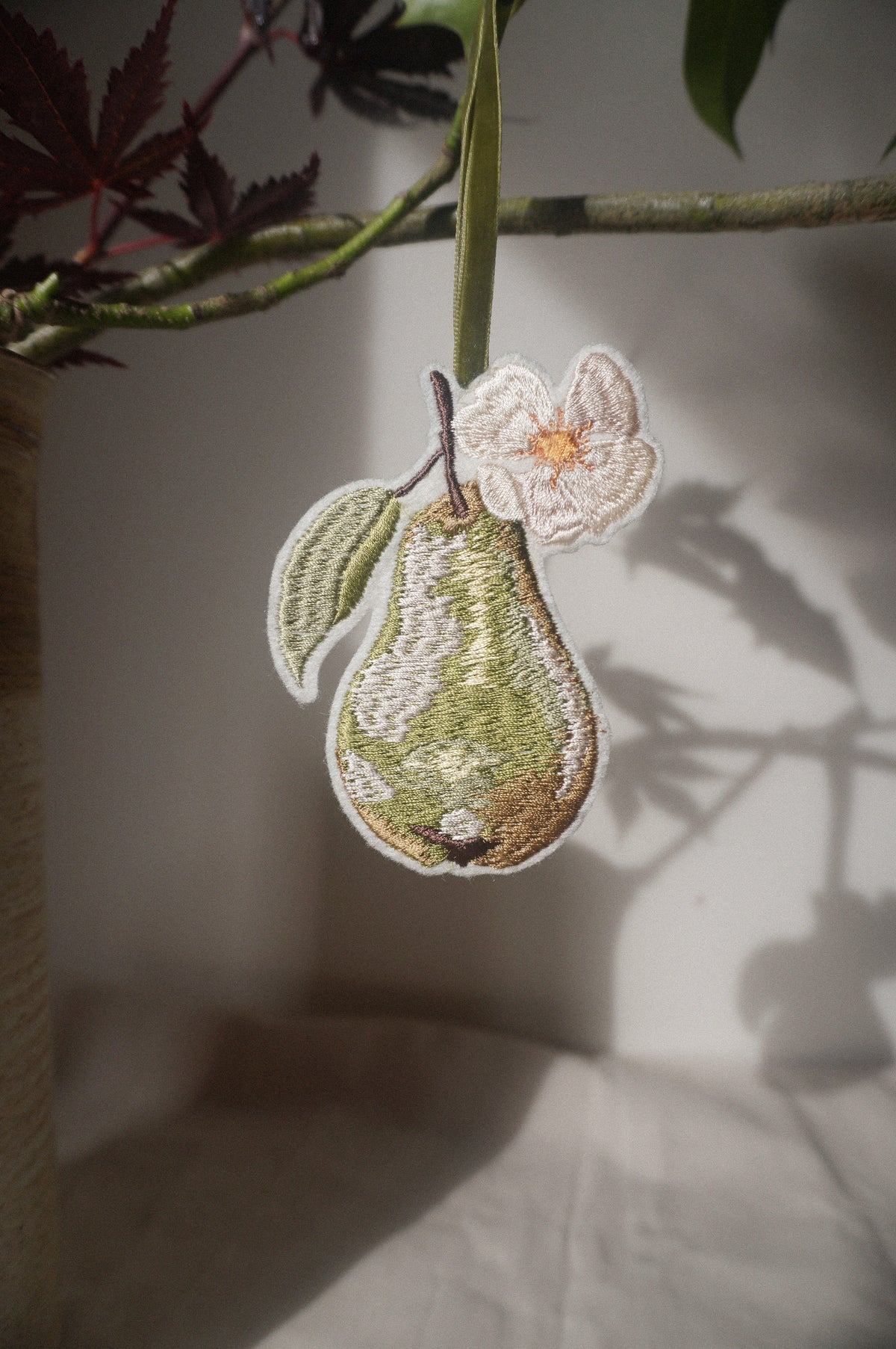 Embroidered green pear decoration with delicate white blossom and leaf, hanging from an olive green velvet ribbon.