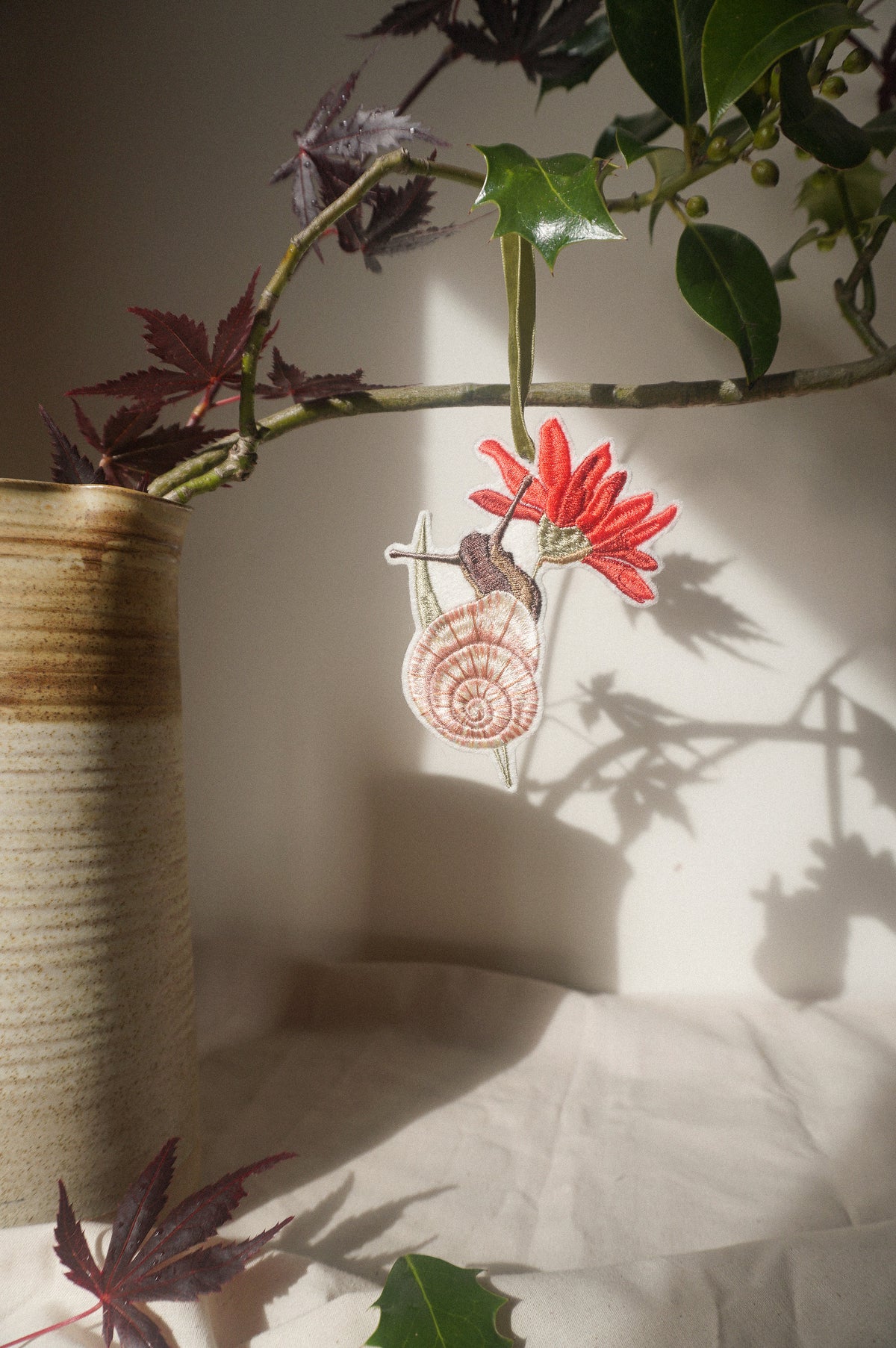 Embroidered garden snail decoration with a pink shell and red flower, hanging from an olive green velvet ribbon amongst leafy branches.