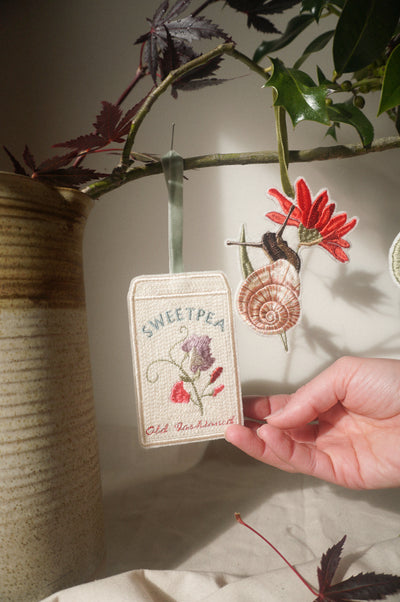 Hand holding an embroidered sweet pea seed packet decoration beside a floral snail ornament, hanging from leafy branches.