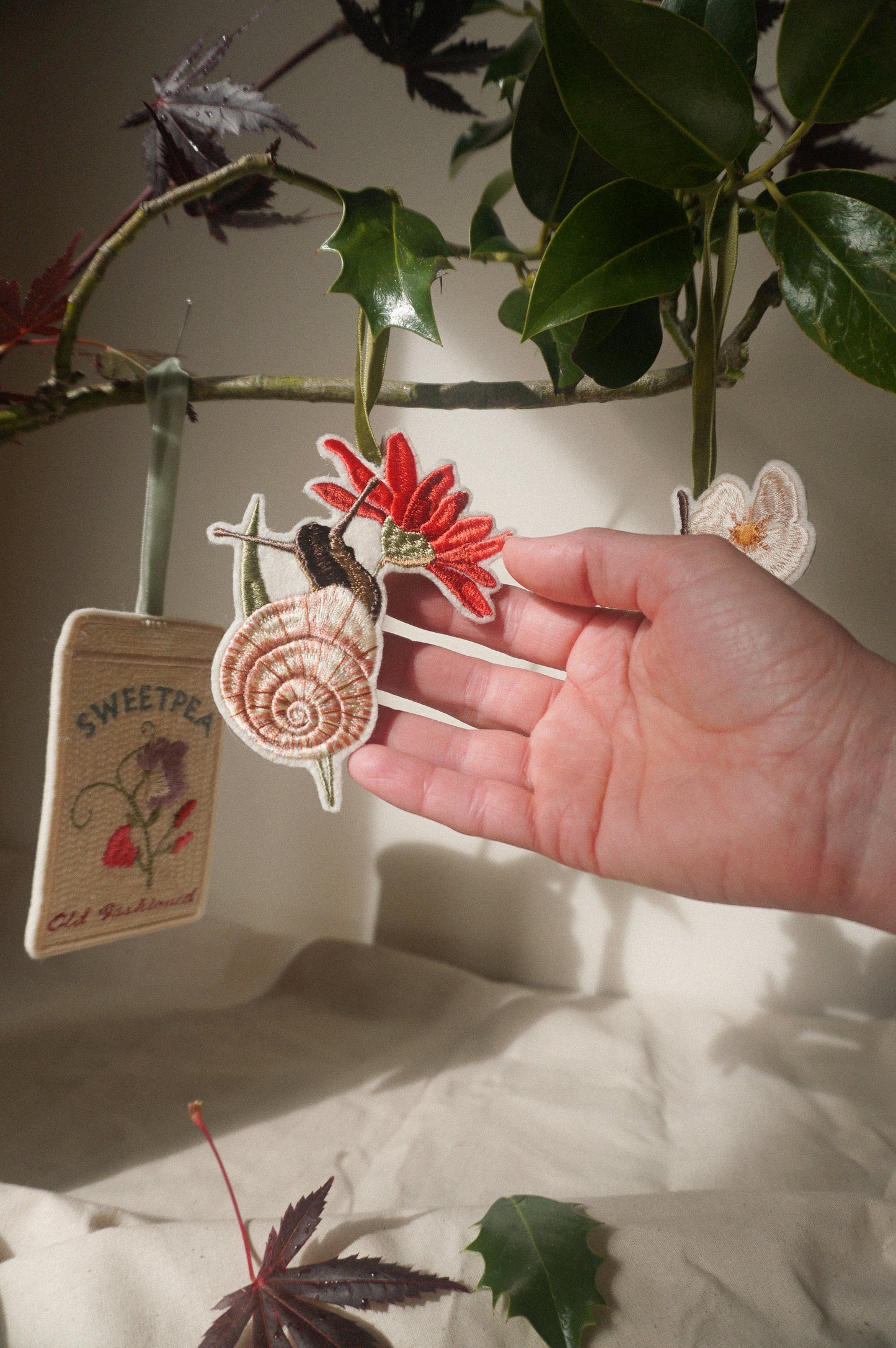 Hand holding an embroidered garden snail decoration with a pink shell and red flower, hanging amongst leafy branches.