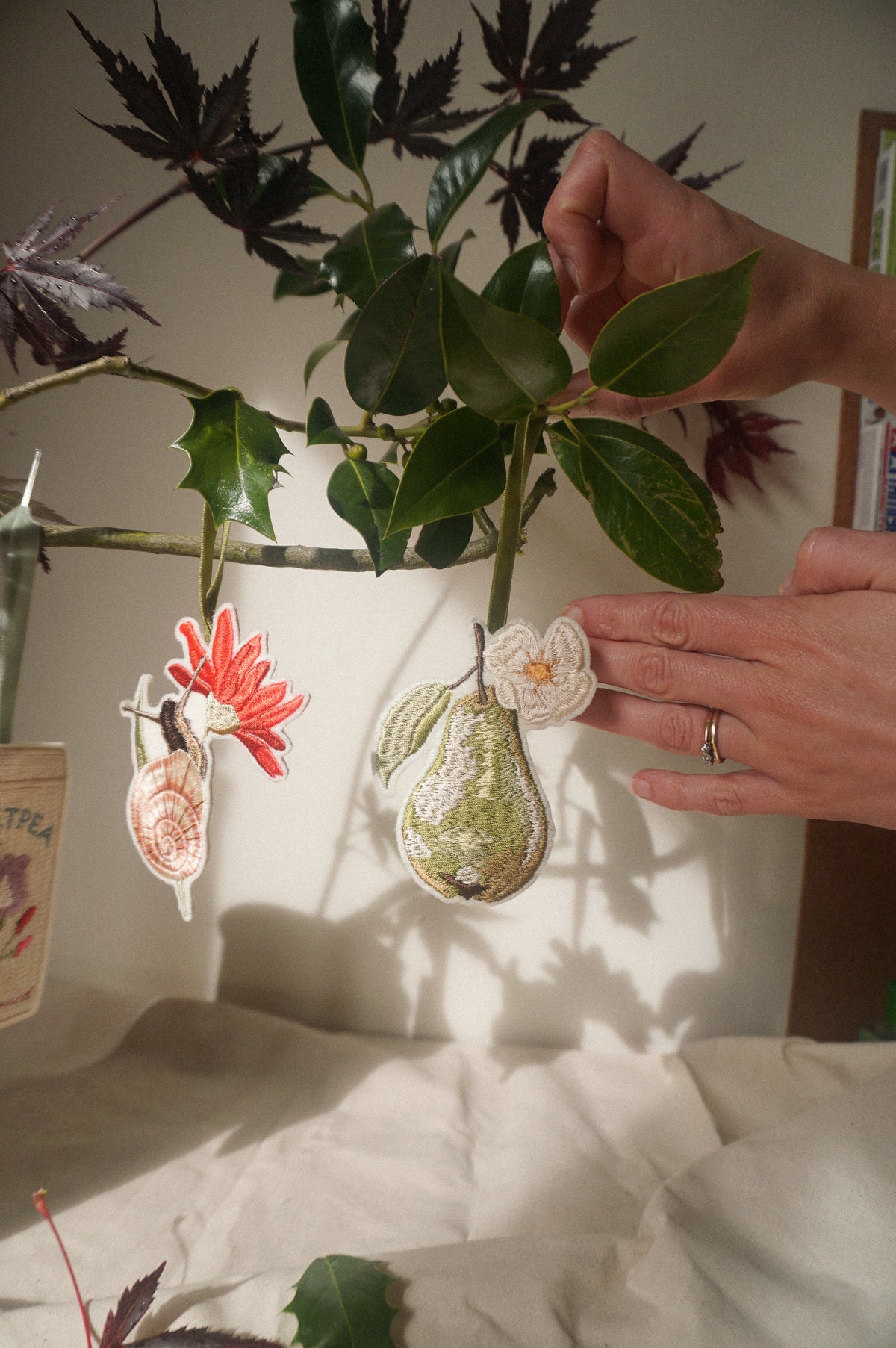 Hands arranging an embroidered pear decoration with blossom beside a garden snail decoration, hanging amongst leafy branches.
