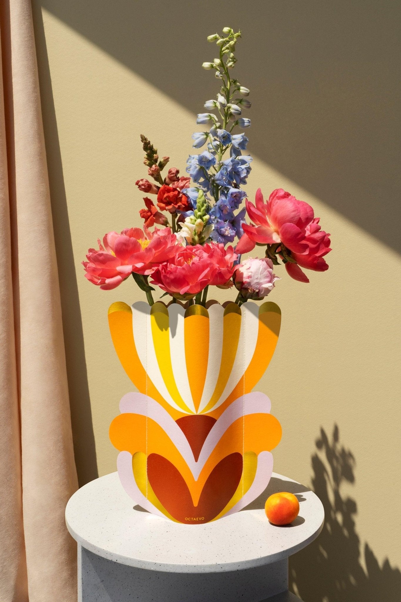 An OCTAEVO Elysia Paper Vase with pink and blue flowers sits on a round white table beside a small orange fruit.