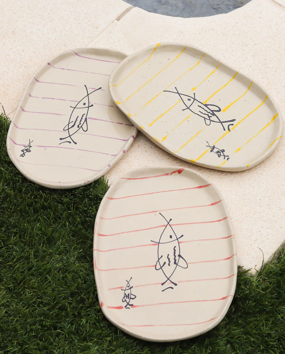 Three ceramic plates with line patterns and fish illustrations on a grassy surface.