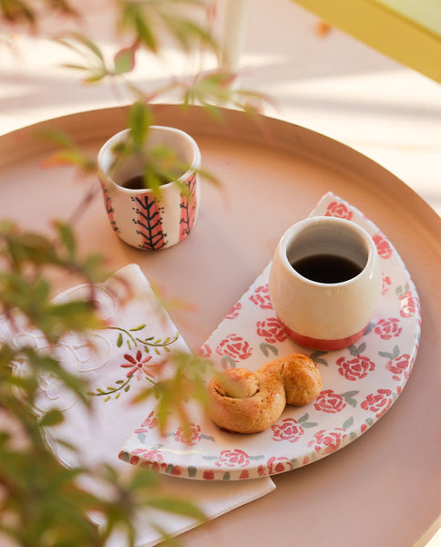 Two ceramic espresso cups with floral patterns, one with a pink base and the other with a white base, placed on a round tray with a matching design, accompanied by a white plate with a rose design, all set on a table with a blurred background.